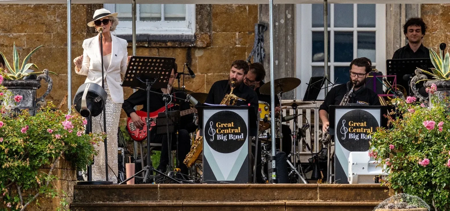 The band playing on the terrace of a stone country house, a singer in a white suit and hat at the microphone, saxophones and guitar behind Great Central Big Band music stands, roses in the foreground